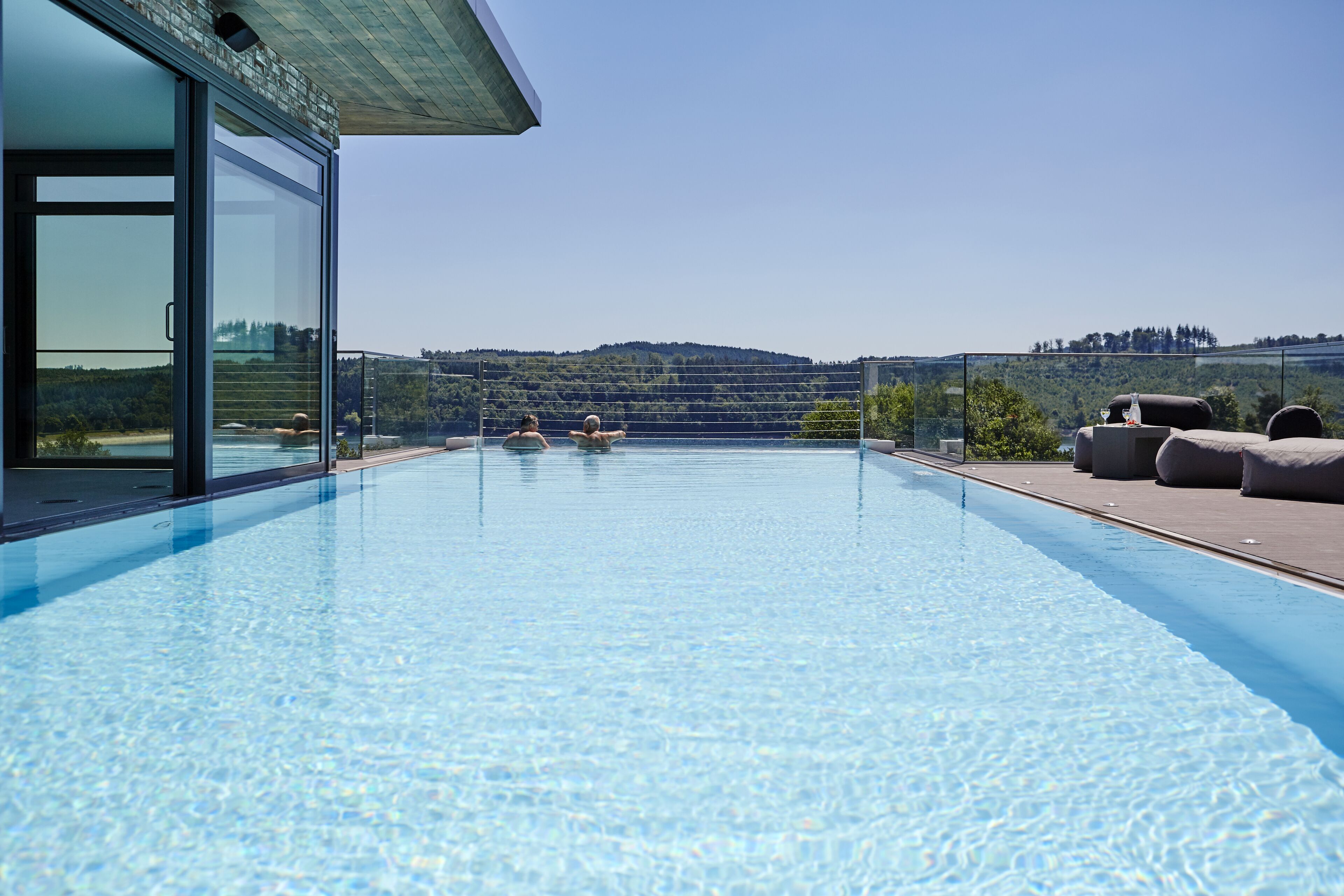 A modern infinity pool with clear blue water, surrounded by a glass enclosure and a wooden deck.