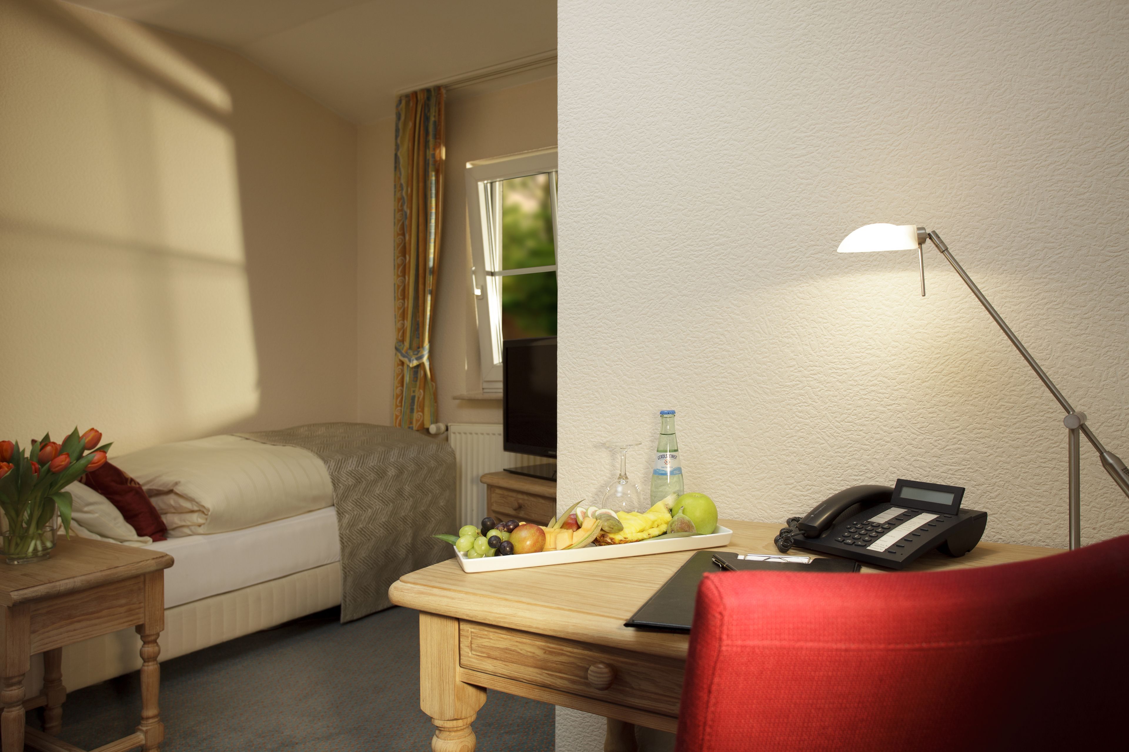 Et hotelværelse med en seng, et skrivebord med lampe, en telefon og et fad med frugt.