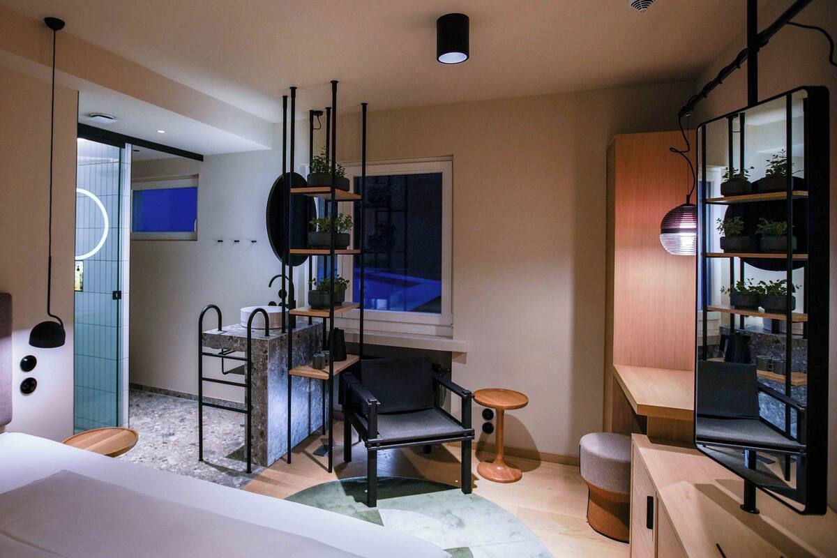 A modern hotel room with a bed, a desk, a chair, a small table, and a mirror. There is a window with a view of the outside.