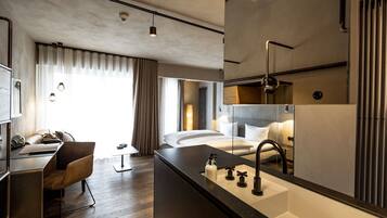 A modern hotel room with a wooden desk, a comfortable chair, a large bed, a bathroom with a mirror, and a view of the city.