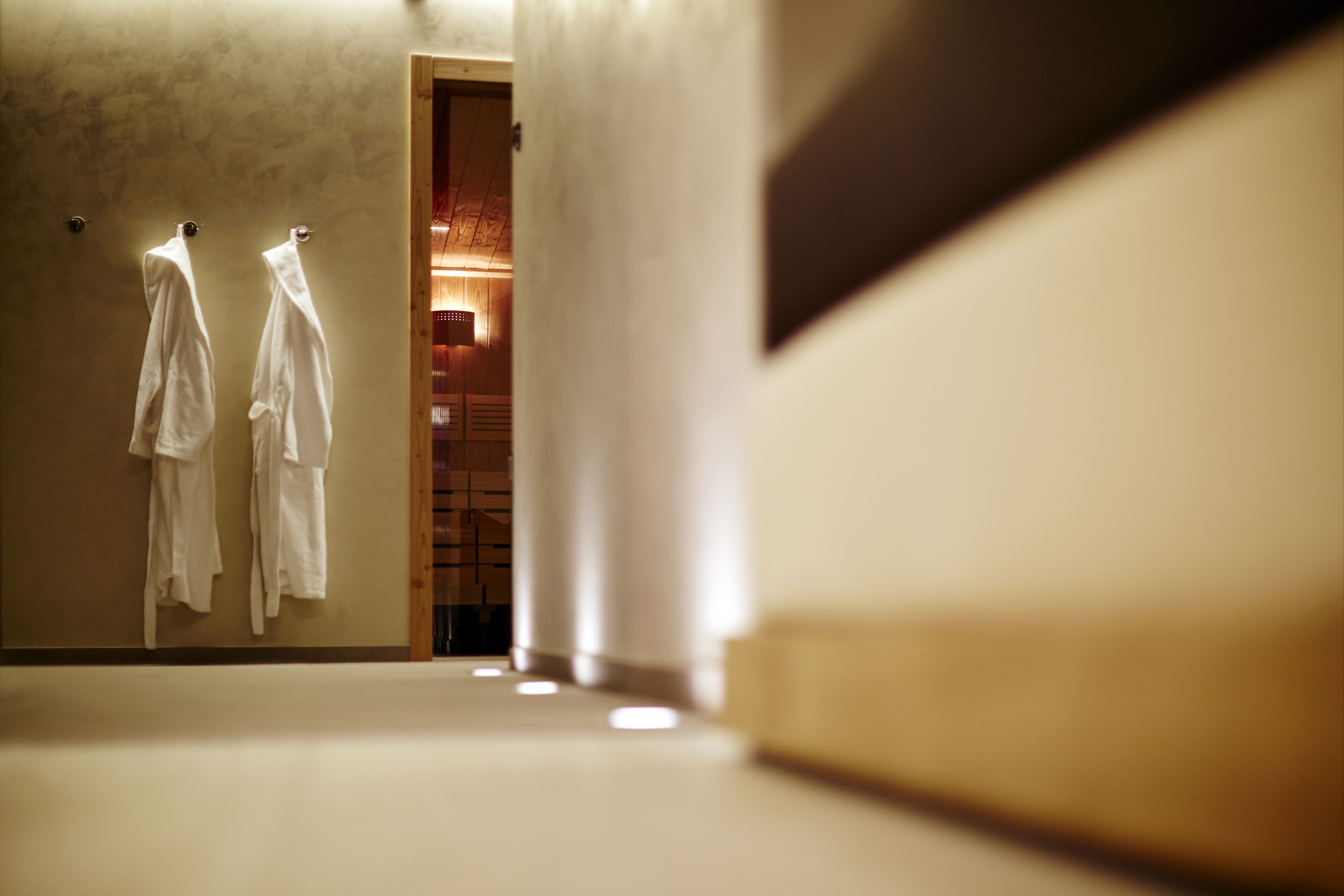 A hallway with two white robes hanging on hooks, leading to a room with wooden paneling and a warm light source.