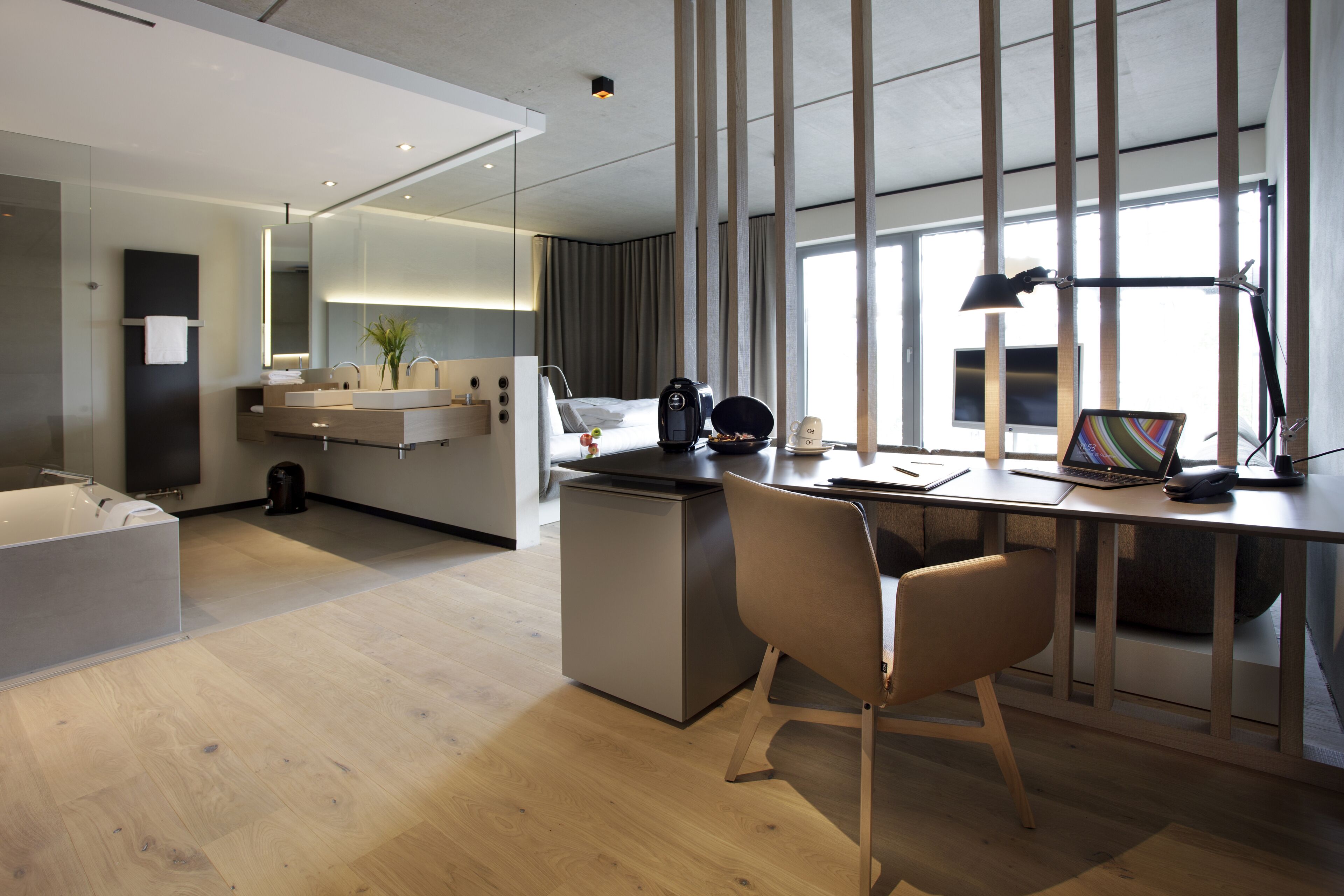 A modern hotel room with a wooden floor, a desk with a laptop, a chair, a coffee maker, and a large window with curtains.