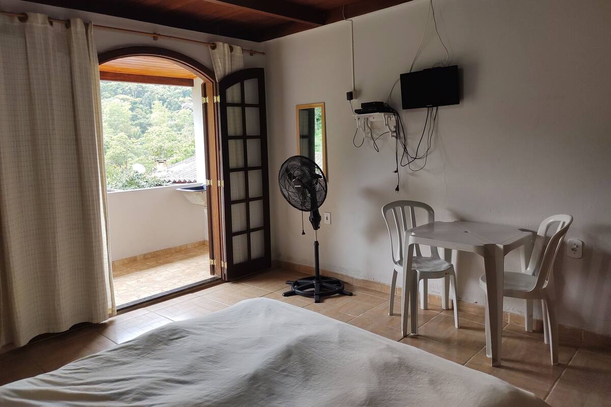 A room with a bed, a table with chairs, a fan, and a TV mounted on the wall.