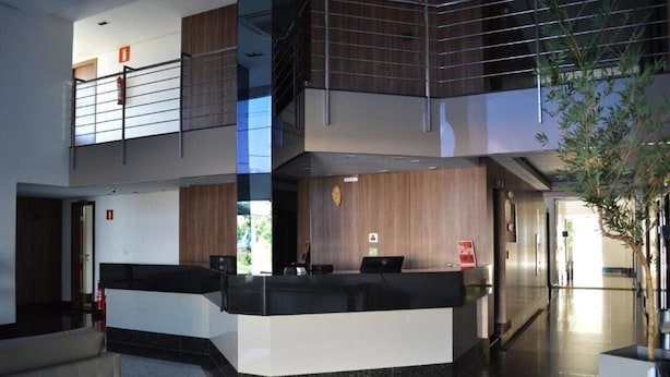 A modern reception area with a black and white counter, a wooden accent wall, and a staircase leading to an upper level. - Liber Hotel Nova Serrana (Nova Serrana)