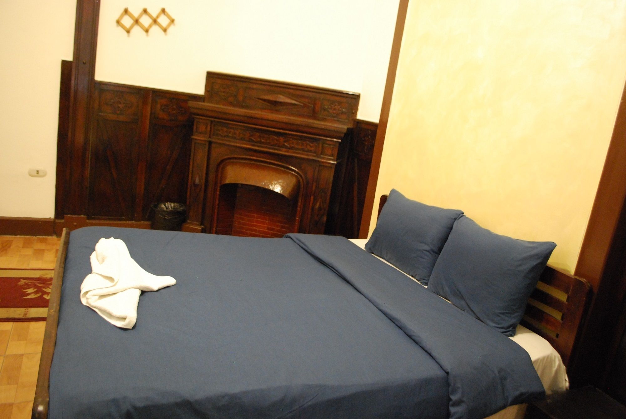 A neatly made bed with a blue bedspread and pillows, a wooden headboard, and a fireplace in the background.