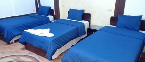 A hotel room with three single beds, each with a blue bedspread and white pillows. There is a small table between the beds, and a decorative rug on the floor.