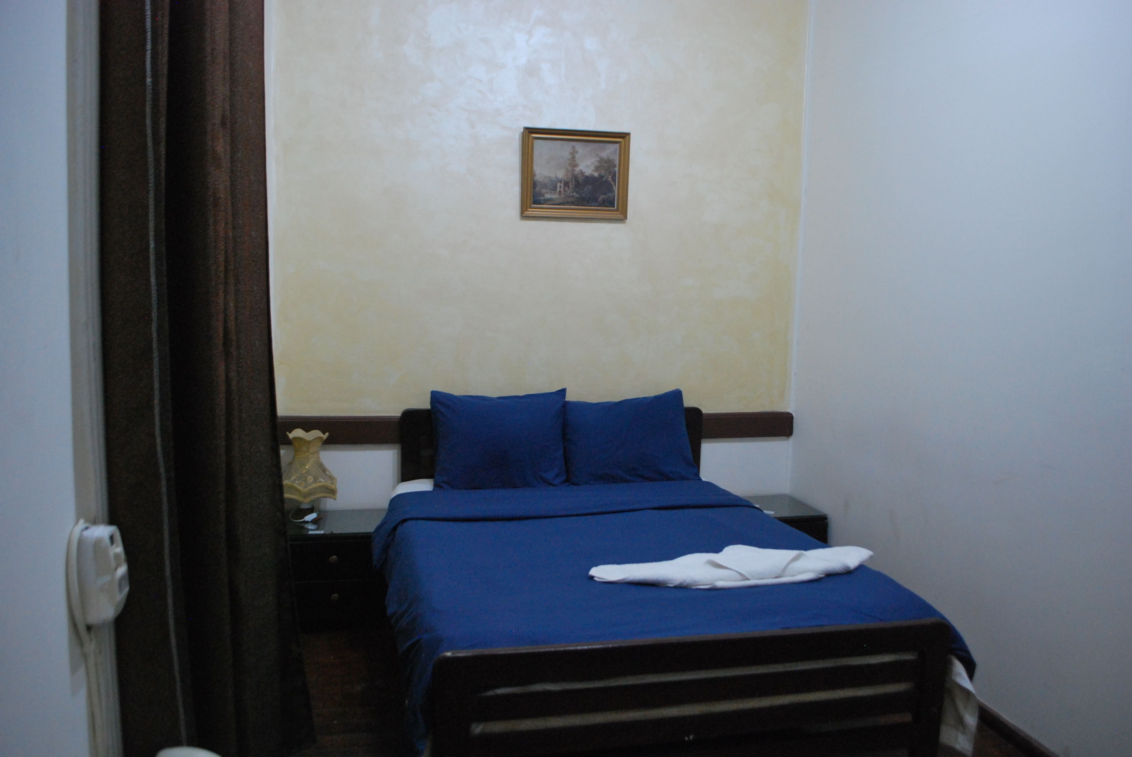Una cama pequeña y ordenada con ropa de cama azul y una toalla blanca encima, una mesita de noche con una lámpara y un cuadro en la pared.