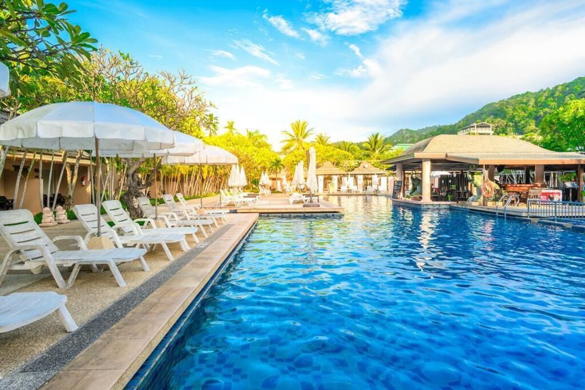 Piscina con sillas de descanso y sombrillas, rodeada de vegetación y un cielo despejado.
