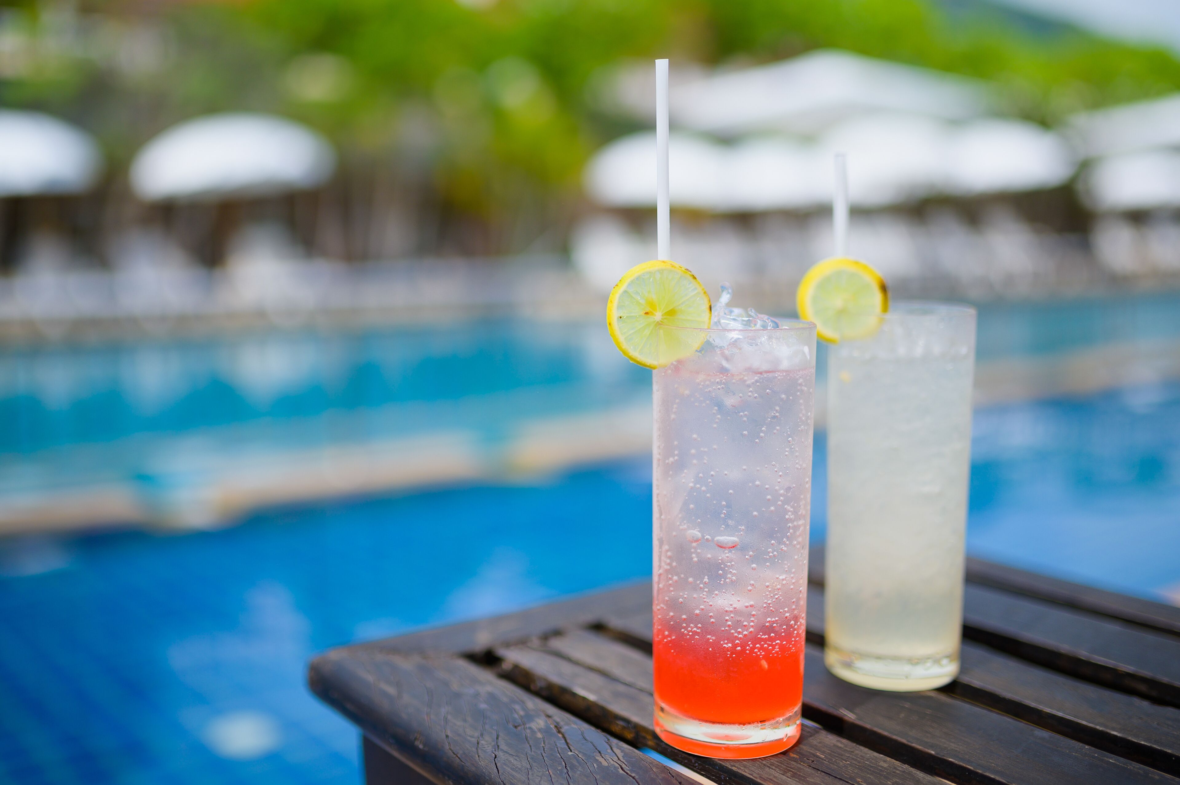 Two glasses of beverages with straws and lemon slices on a wooden table by a pool.