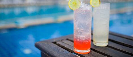 Two glasses of beverages with straws and lemon slices on a wooden table by a pool.