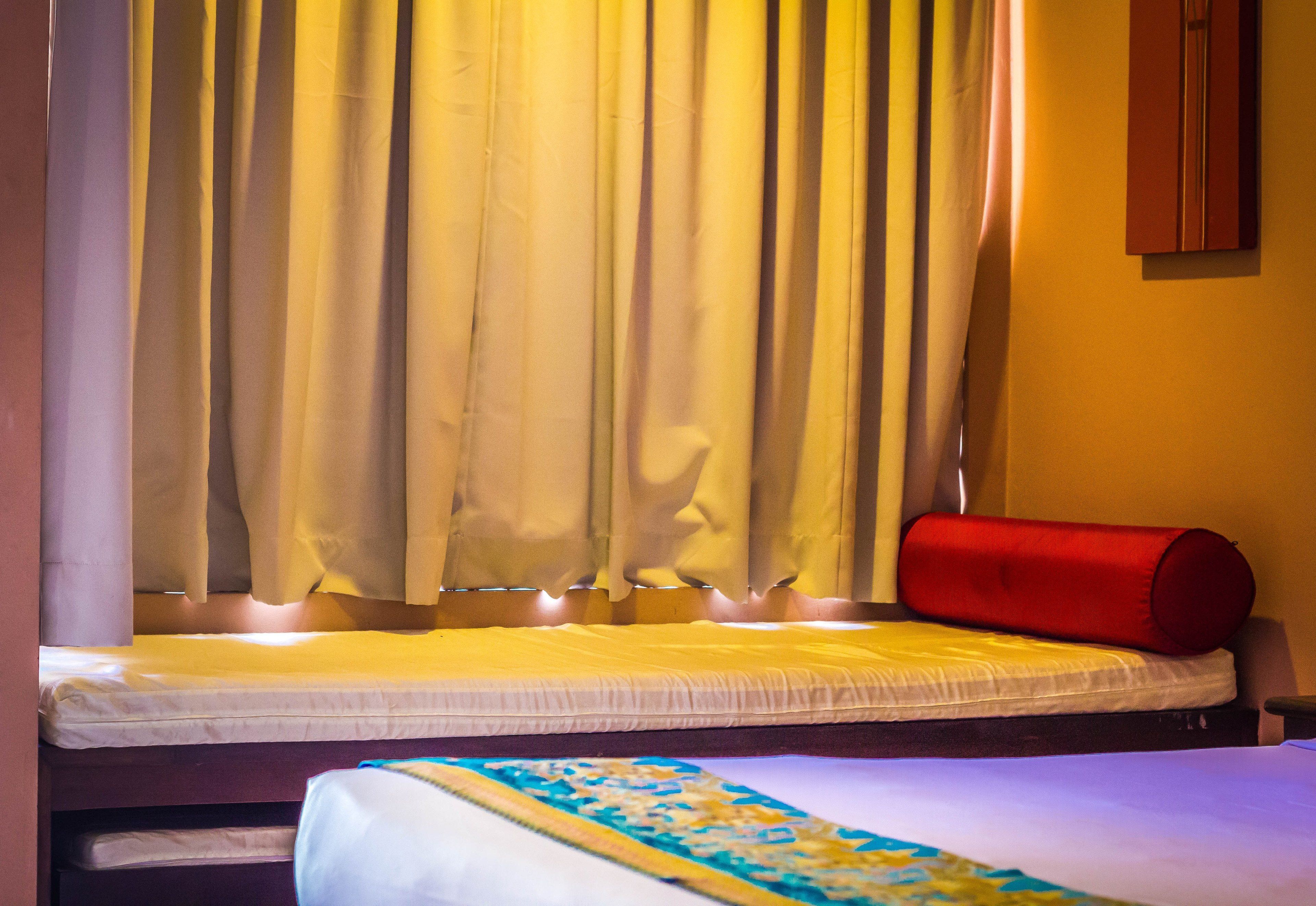 A single bed with a white mattress and a red pillow, a yellow blanket, and a window with yellow curtains.