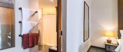 A hotel bathroom with a shower, a towel rack with towels, a small table with a lamp, and a framed picture on the wall.
