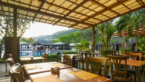 An outdoor restaurant with wooden tables and chairs, a pool in the background, and lush greenery.