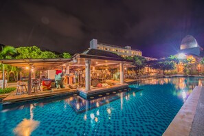 Piscina con área techada y sillas reclinables, rodeada de abundante vegetación y un edificio con techo abovedado.