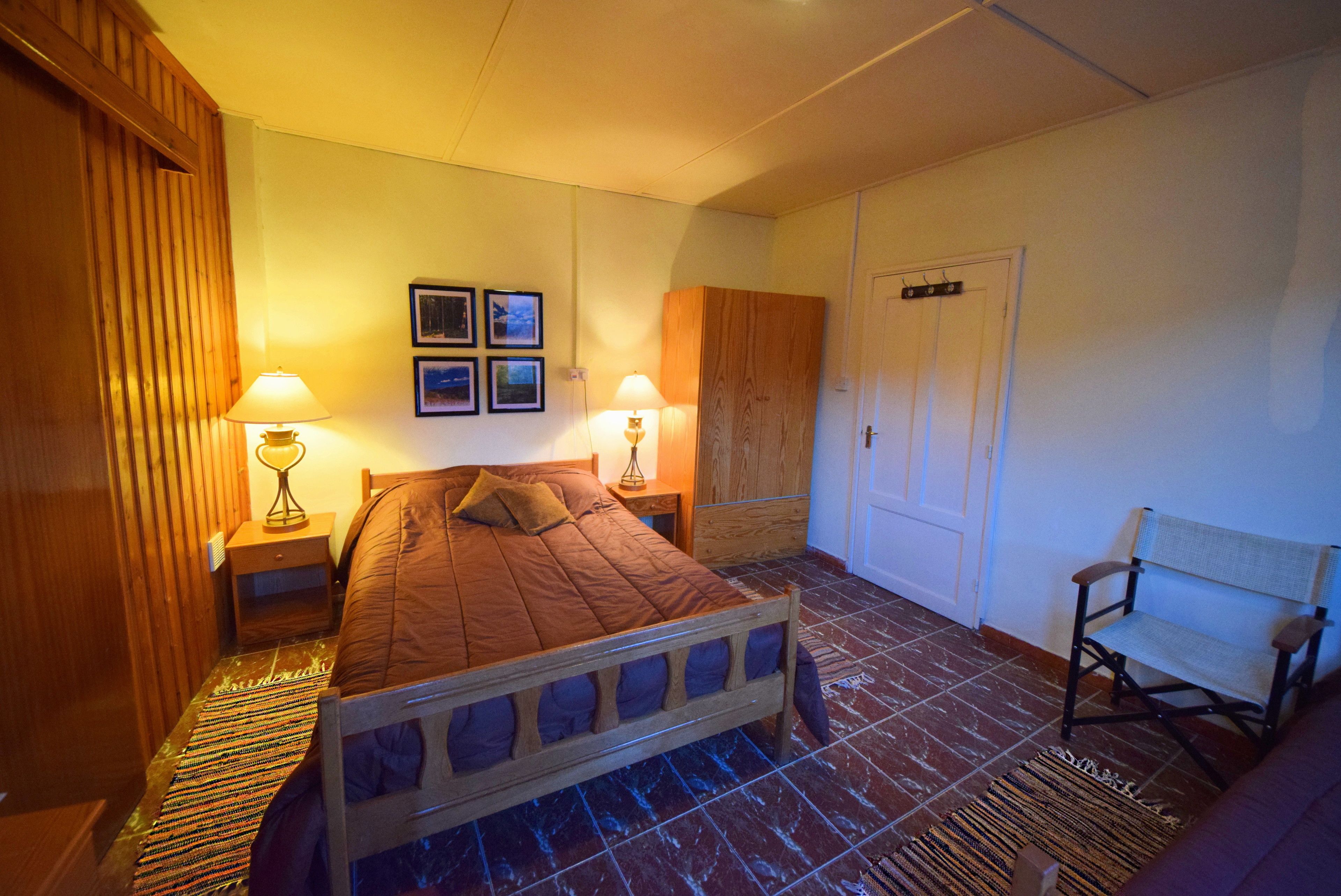 A bedroom with a wooden bed, two bedside lamps, a wooden wardrobe, a chair, and a door.