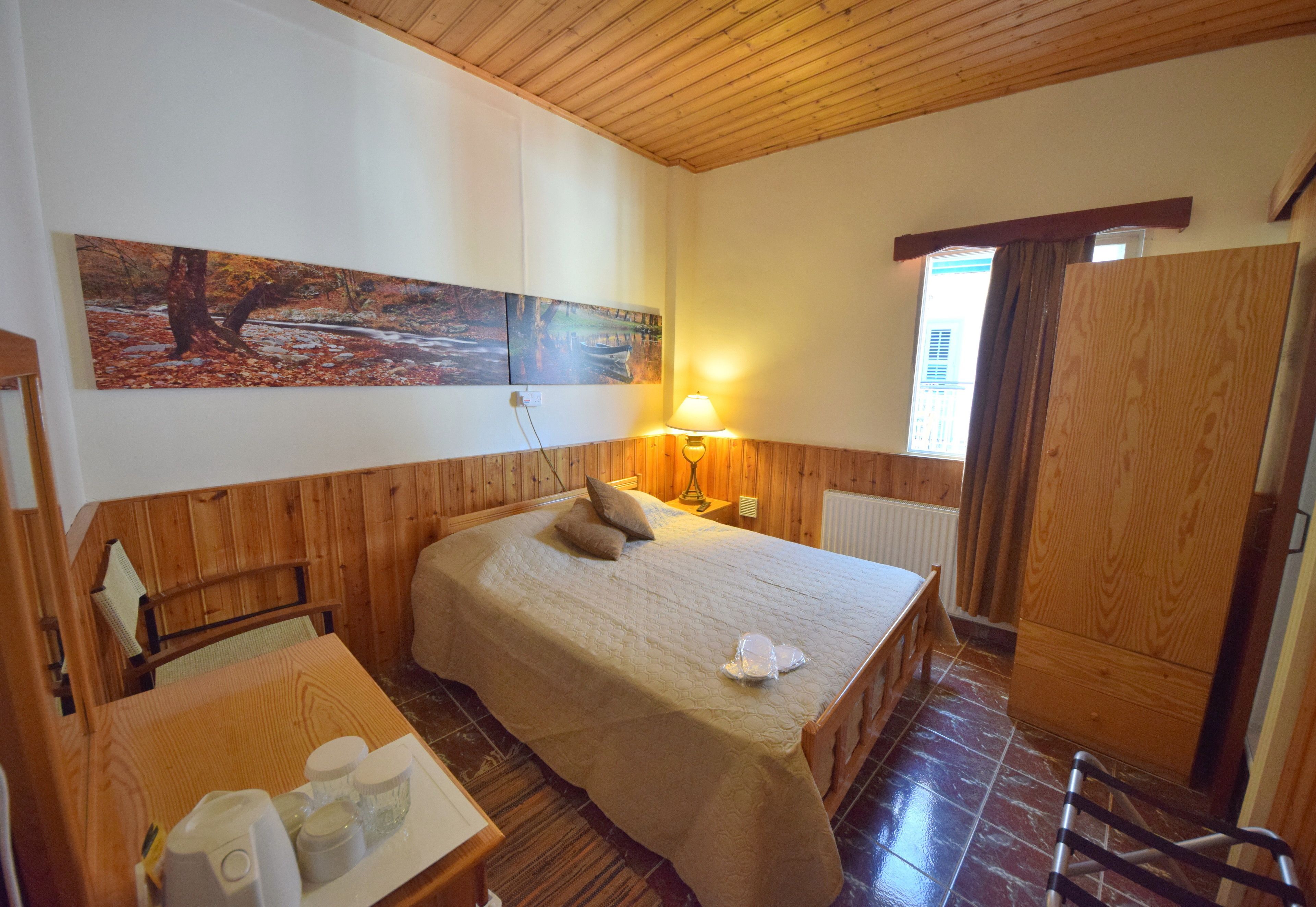 Una habitación con paneles de madera, con una cama, una mesita de noche con lámpara, un radiador y una ventana con cortinas.