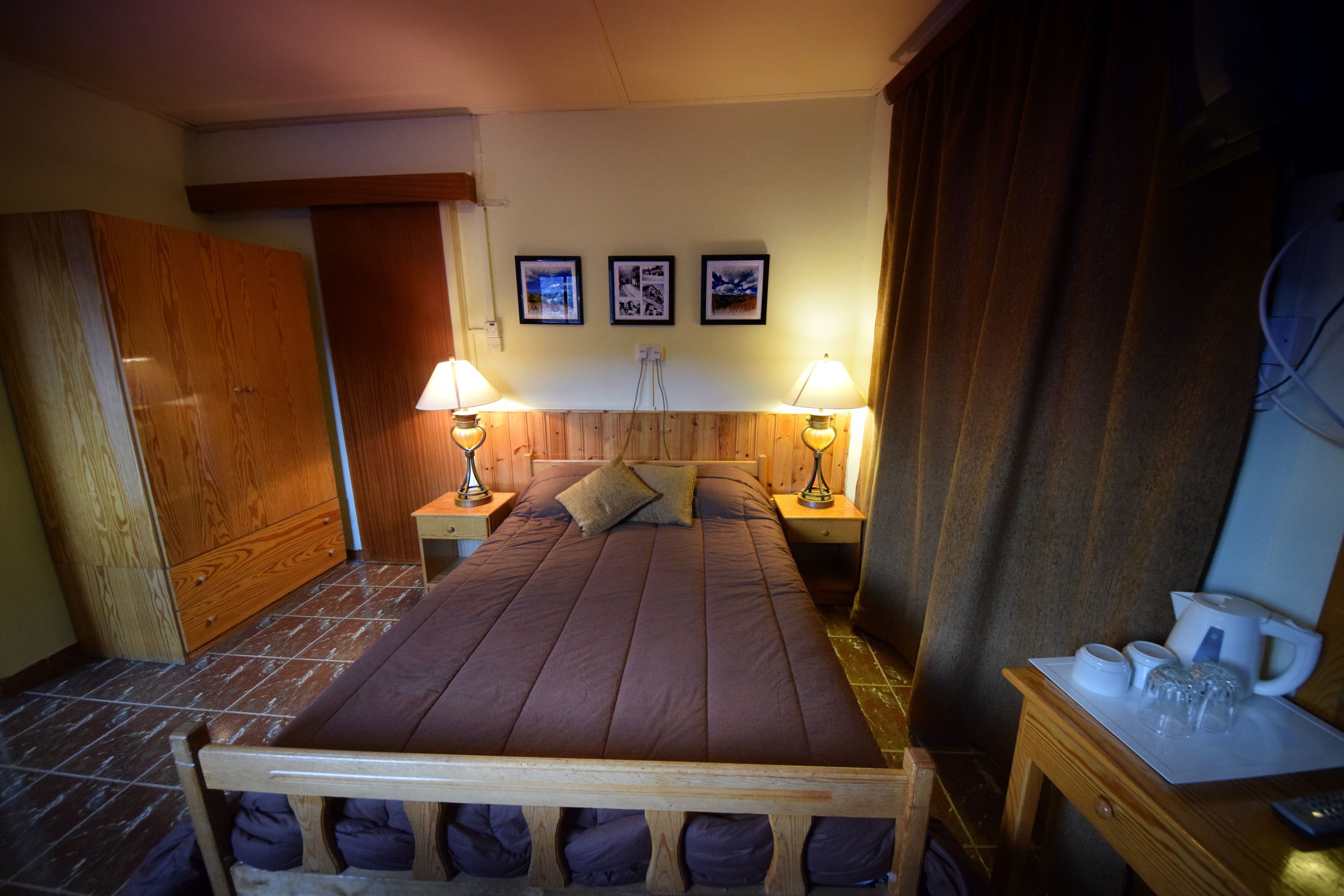 Un dormitorio con una cama de madera, dos lámparas de noche, una mesita de noche con cafetera y un armario de madera.