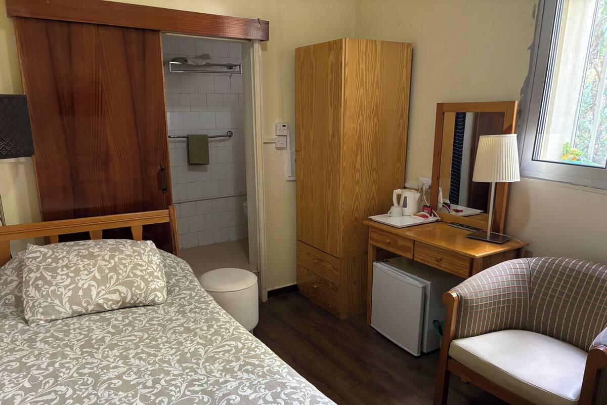 Een knus hotelkamer met een bed, een bureau met stoel, een kleine koelkast, een houten garderobe en een douchehoek.