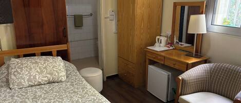 Une chambre d’hôtel compacte comprenant un lit, un bureau avec une chaise, un petit réfrigérateur, une armoire en bois et un espace douche.