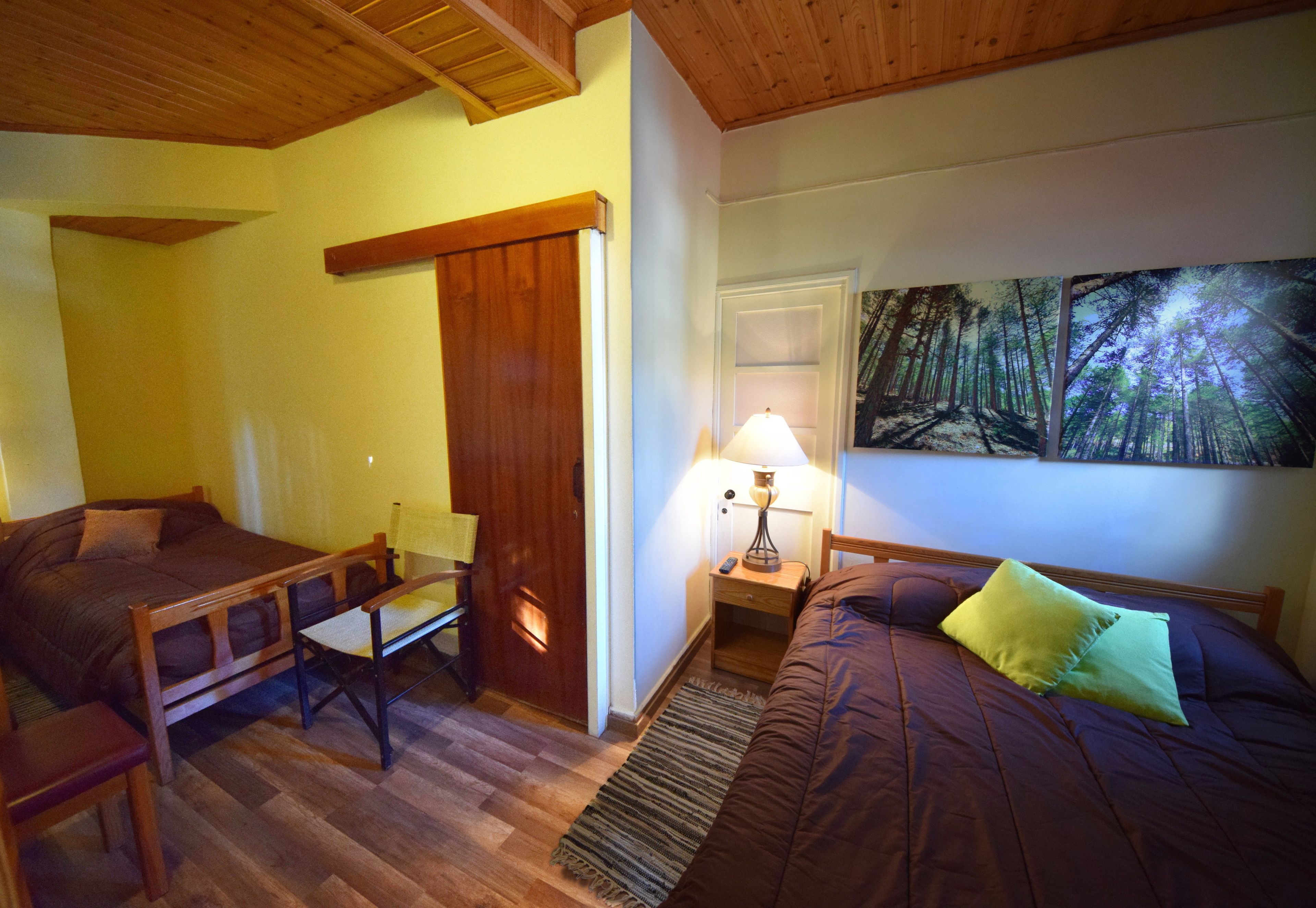 A room with two beds, a chair, a nightstand with a lamp, and a painting of a forest on the wall.