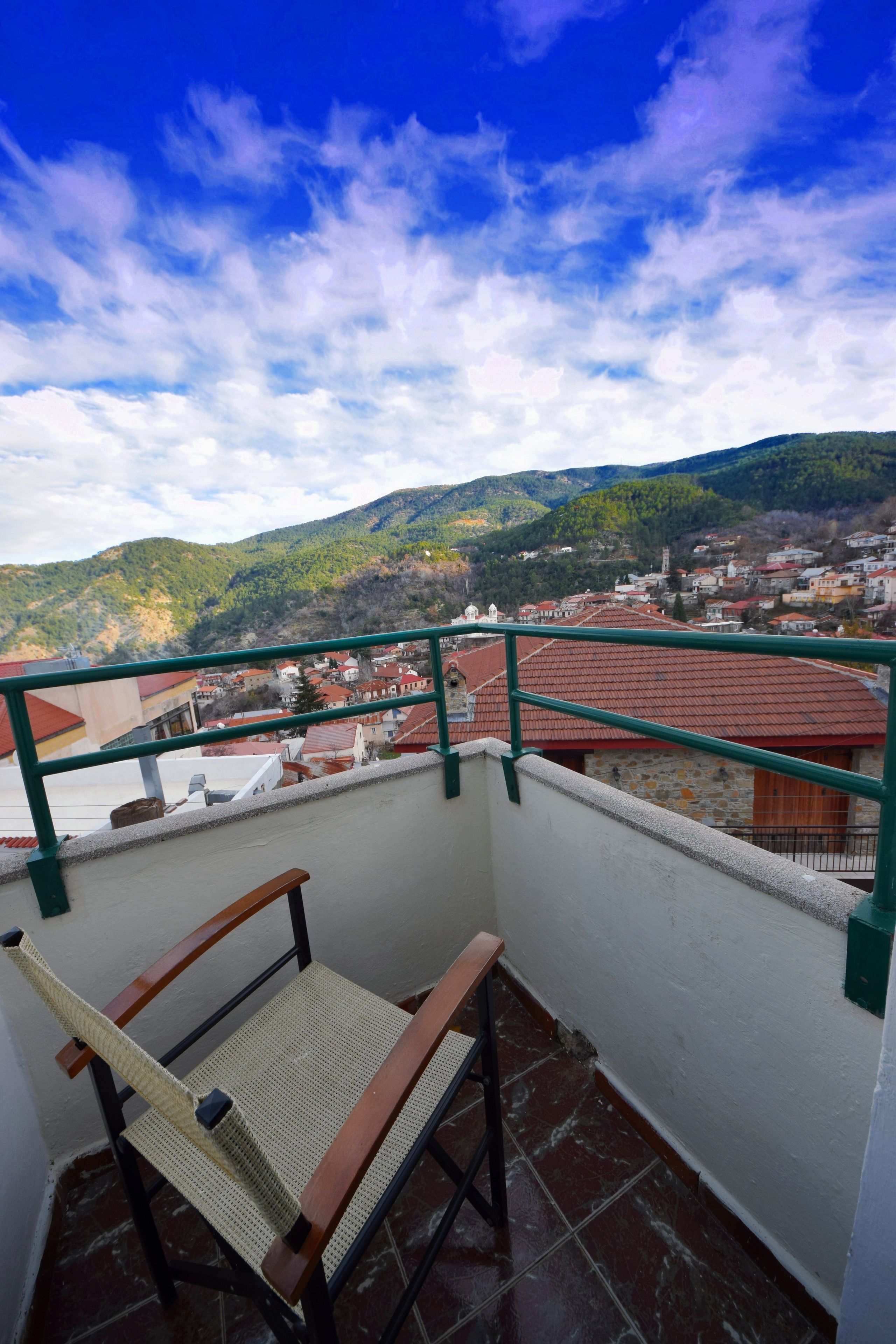 A balcony with a wooden chair and a view of a town with red-roofed buildings and green hills in the background.