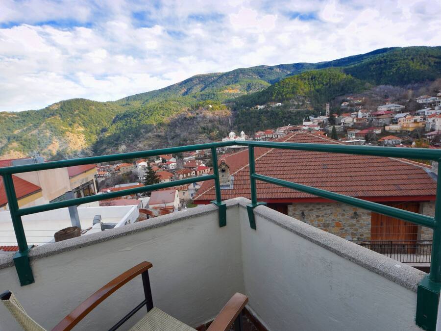 A balcony with a wooden chair and a view of a town with red-roofed buildings and green hills in the background.