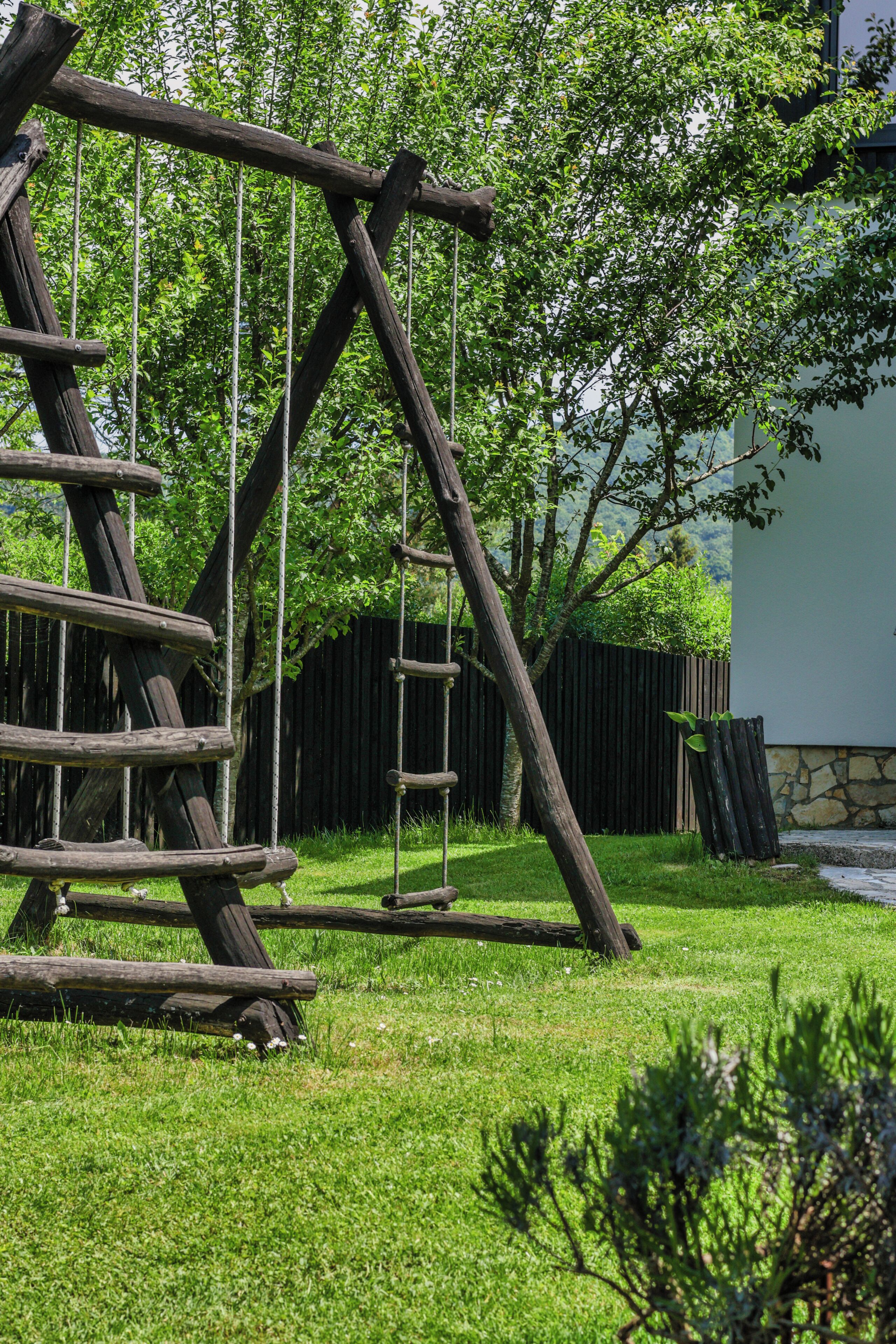 A wooden climbing structure in a grassy area with trees and a stone wall in the background.