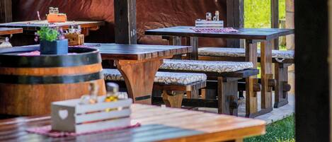 Un área de comedor al aire libre con mesas y bancos de madera, techo cubierto y vistas a zonas verdes.