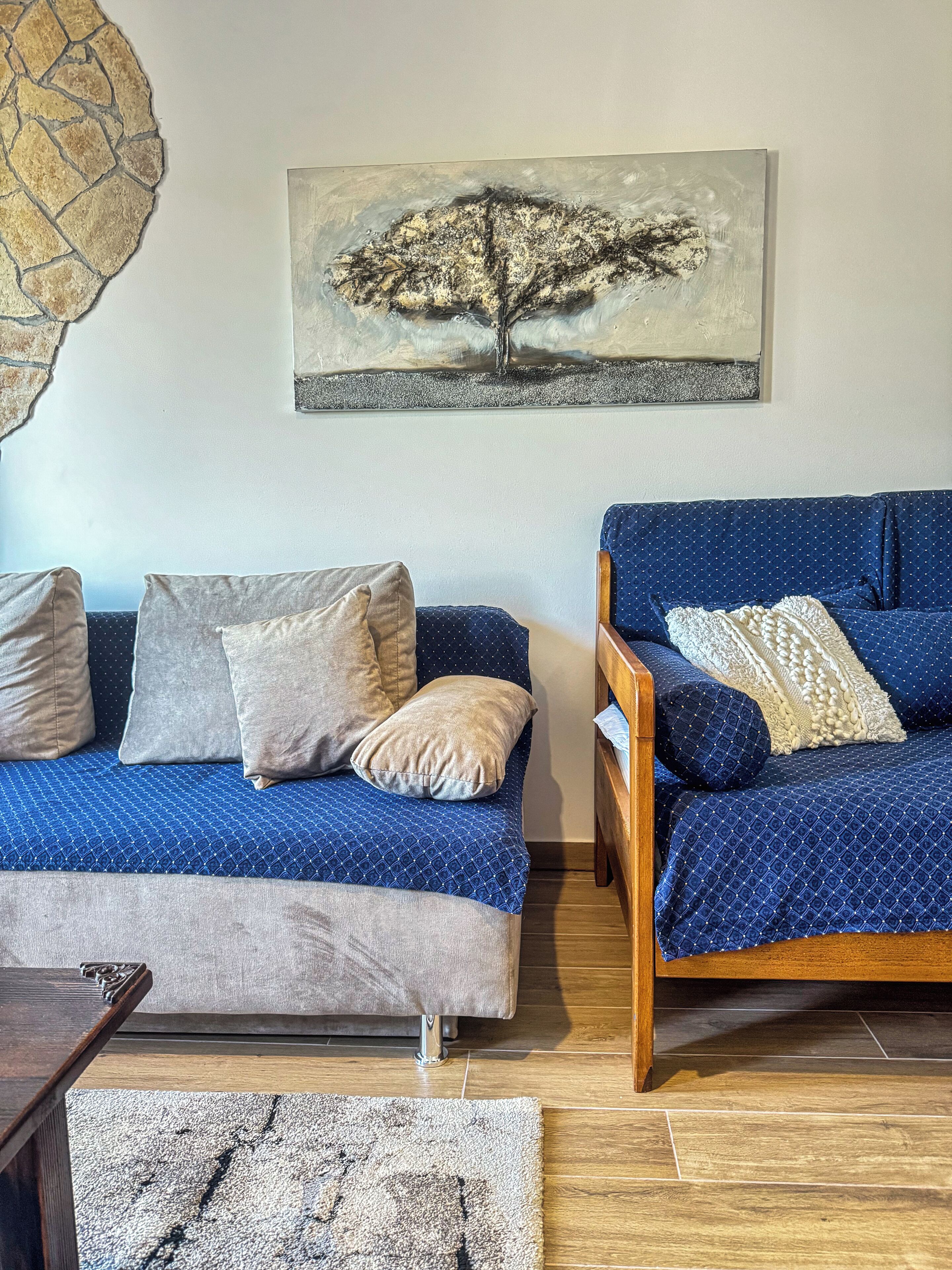Un salon avec un canapé bleu, un fauteuil en bois et un tableau représentant un arbre accroché au mur.