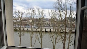 Blick durch ein Fenster auf einen Fluss mit kahl stehenden Bäumen und einer Brücke im Hintergrund.