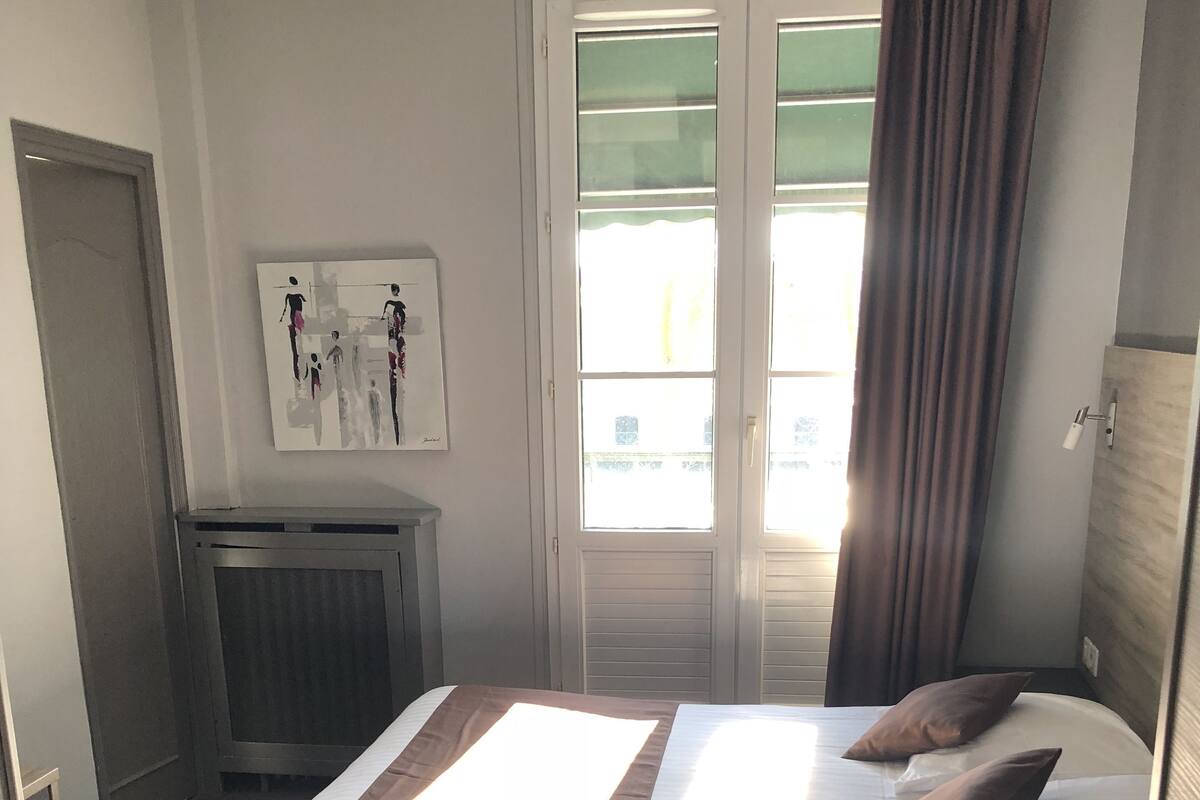 Habitació d'hotel amb un llit, un radiador, una porta, una finestra amb cortines i un quadre penjat a la paret.