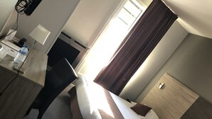 Ein Hotelzimmer mit einem Bett, einem Schreibtisch mit Lampe, einer Wasserflasche und einem Fenster mit Vorhängen.