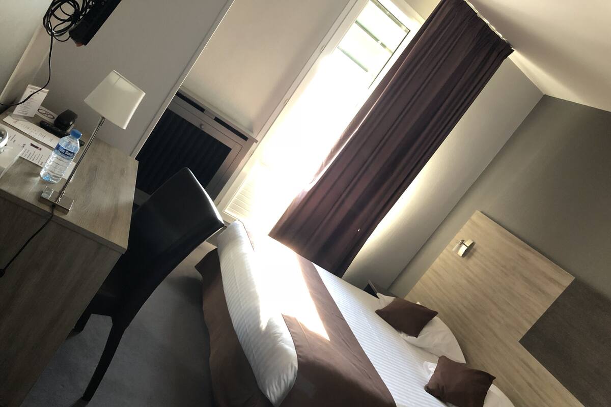 Habitació d'hotel amb llit, escriptori amb làmpada, ampolla d'aigua i finestra amb cortines.