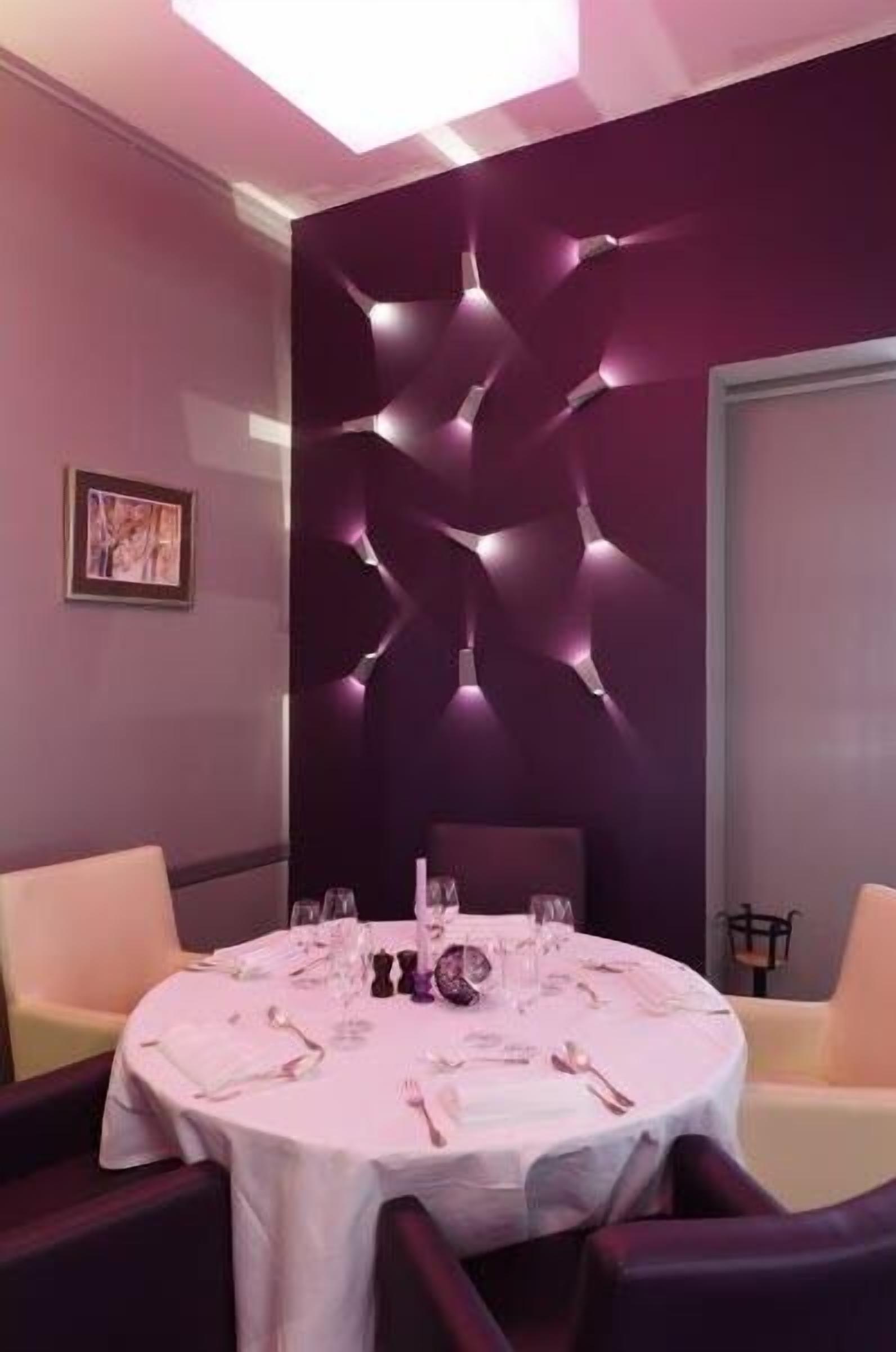 Un coin repas avec une table ronde dressée pour quatre personnes, un mur moderne agrémenté d’un éclairage décoratif abstrait et un tableau encadré accroché au mur.