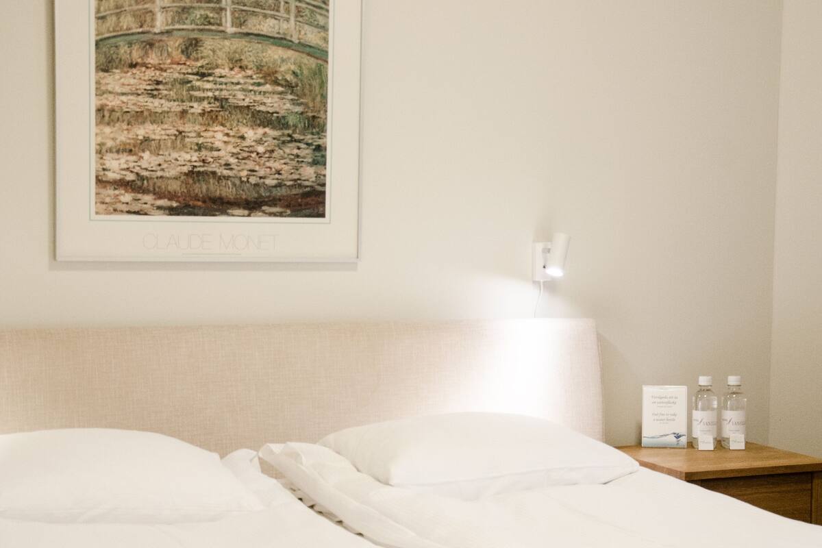 A neatly made bed with a white comforter and a folded towel, a framed painting of a bridge over water, a bedside lamp, and a nightstand with two bottles.