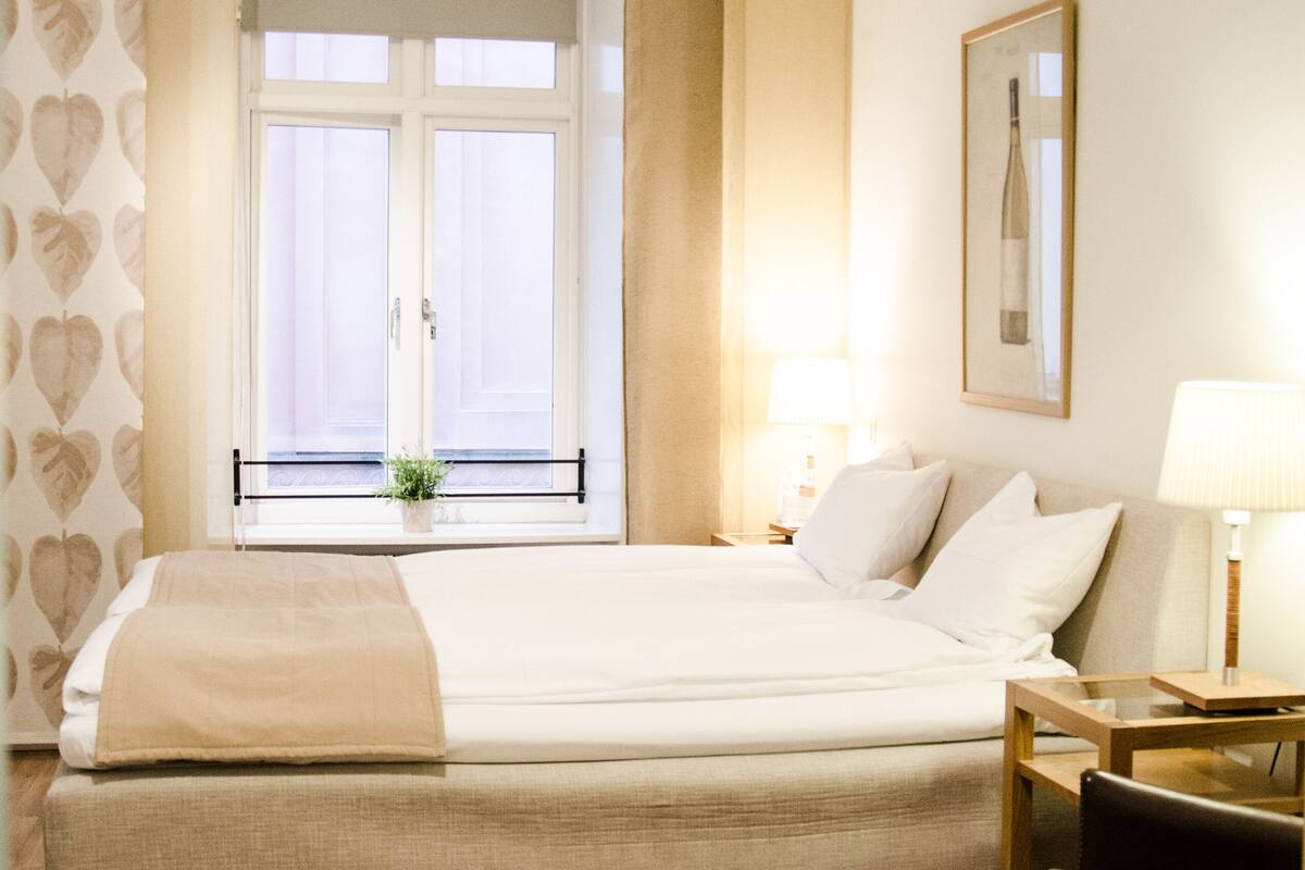 A hotel room with a large bed, a window with curtains, a bedside table with a lamp, and a chair.