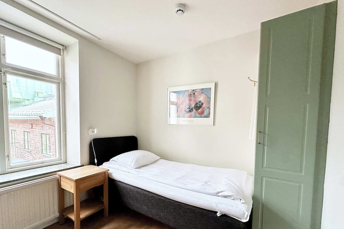 A small, single-bed room with a wooden desk, a window with a view of buildings, and a green door.