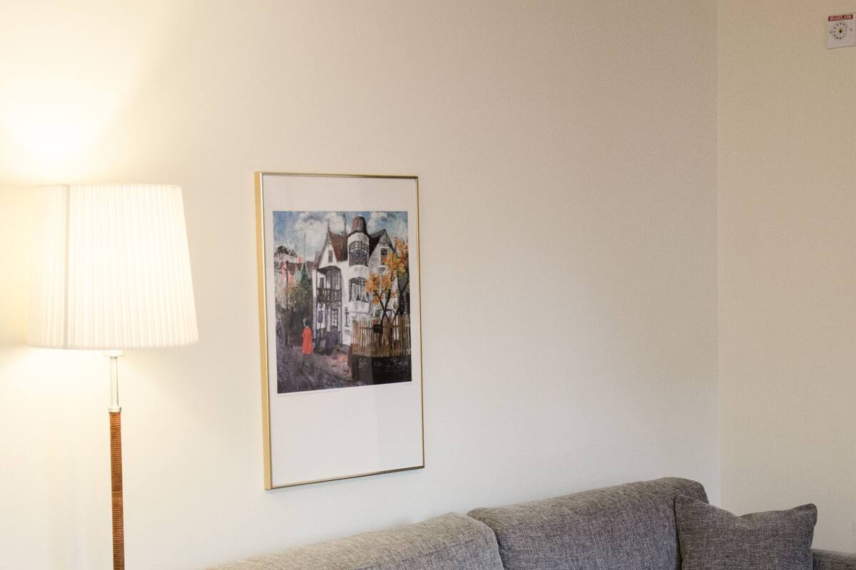 A hotel room with a grey sofa, a white lamp, a framed picture on the wall, and a bed with a white cover.