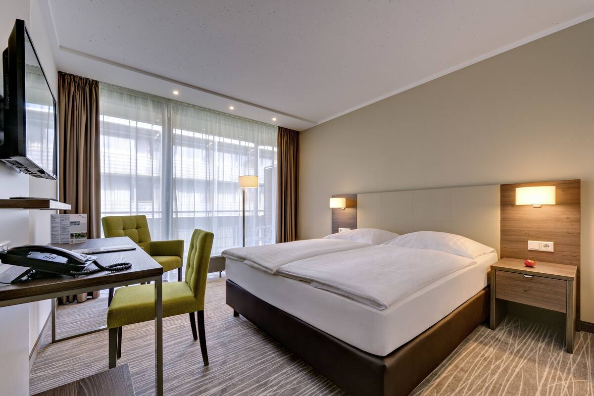 A hotel room with a bed, a desk with a telephone, a chair, a TV, and a lamp.