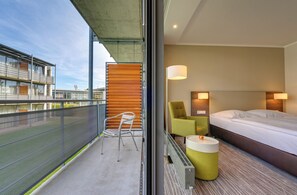 A hotel room with a bed, a chair, a small table, a lamp, and a view of the outside. - GenoHotel Baunatal (Baunatal)