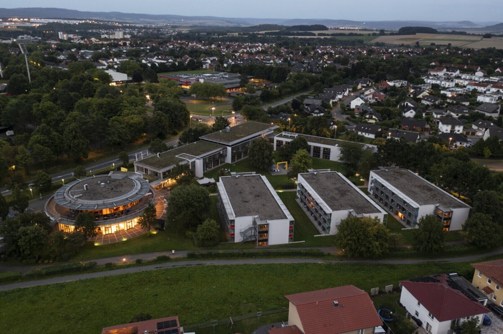 An aerial view of a campus with multiple buildings, including a circular structure with a central courtyard and several rectangular buildings with flat roofs.