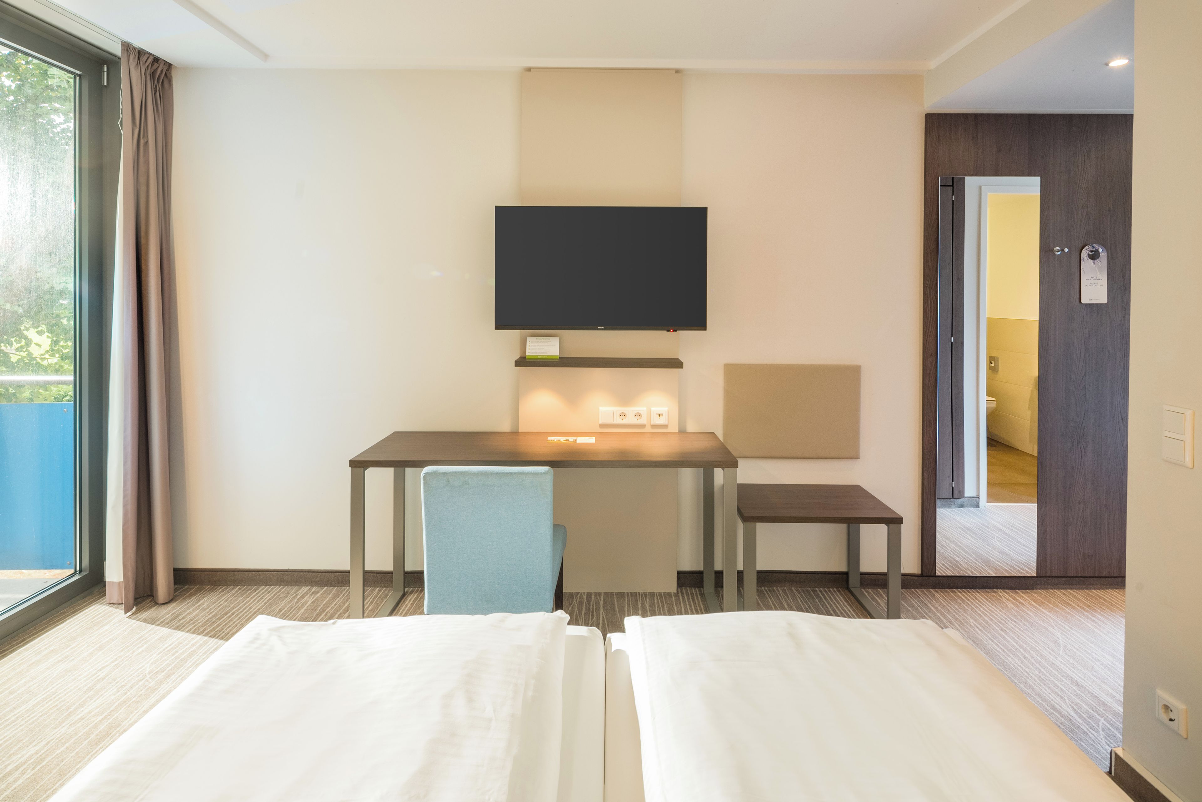 Habitación de hotel con dos camas, un escritorio con silla, televisor de pantalla plana montado en la pared y una puerta que da a otra habitación.