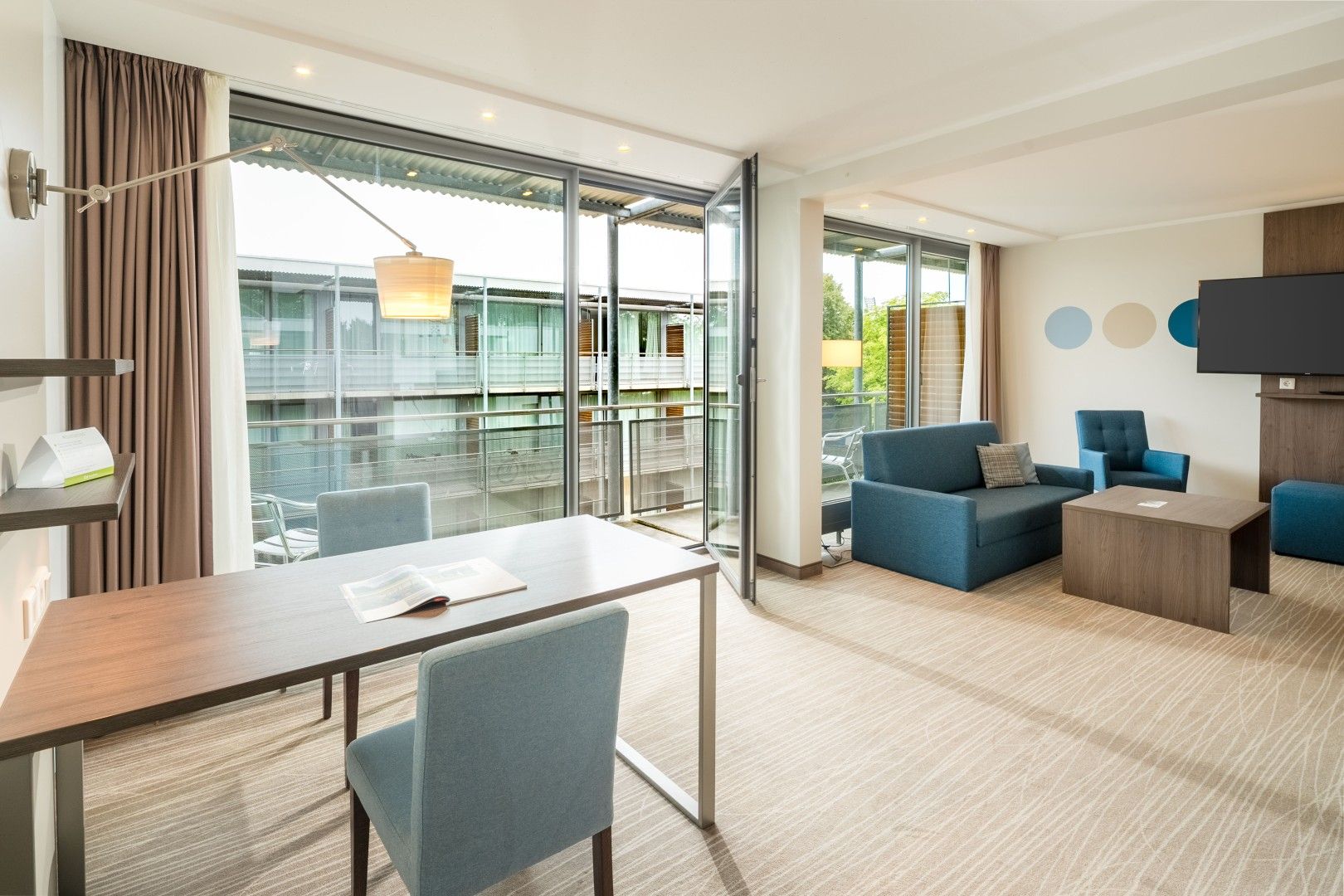 Ein modernes Hotelzimmer mit Essbereich, Sitzgelegenheit und Fernseher, sowie große Fenster mit Blick nach außen.