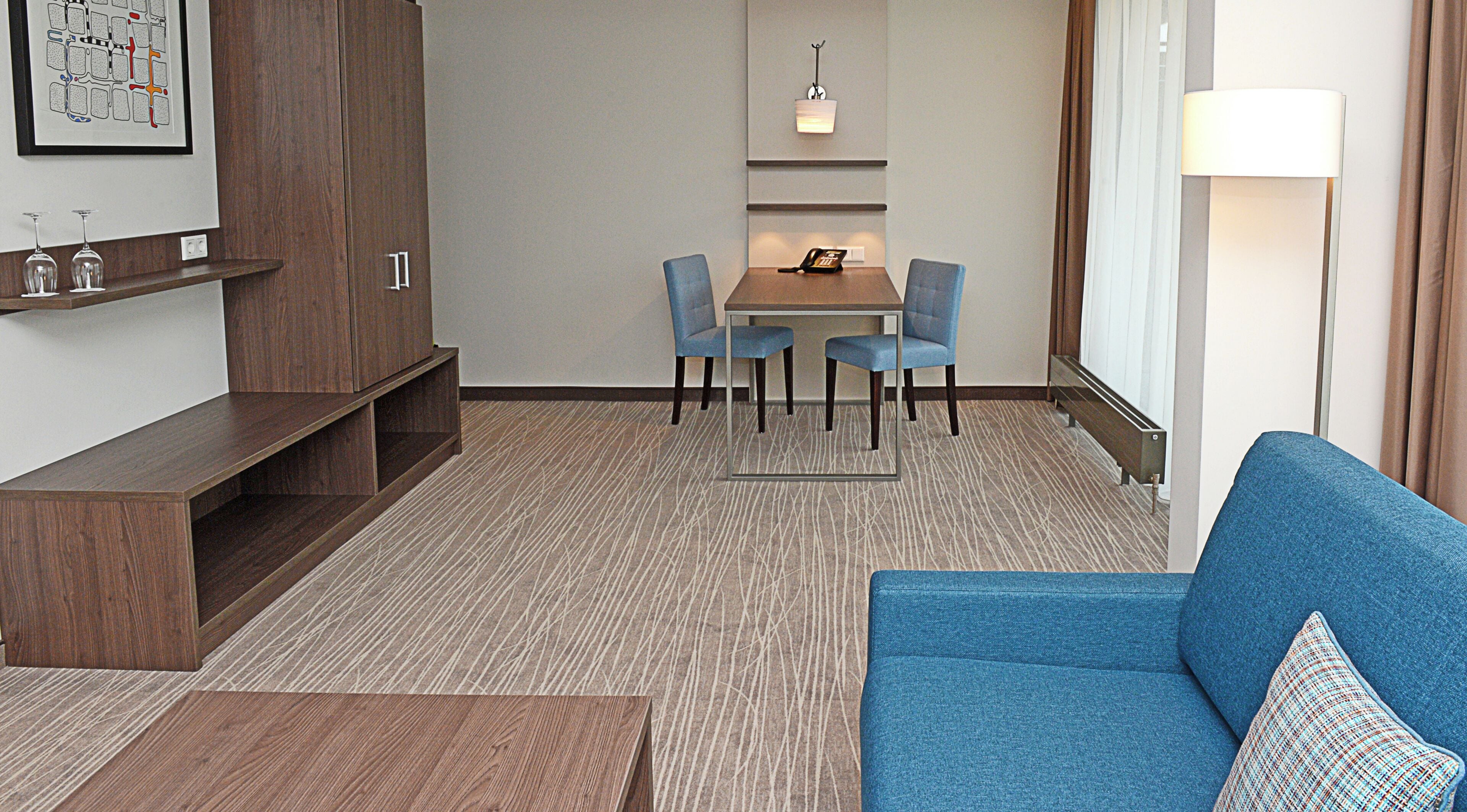 Ein Hotelzimmer mit einem blauen Sofa, einer Holzkassette, einem Esstisch mit Stühlen, einem kleinen Schreibtisch und einem an der Wand befestigten Telefon.