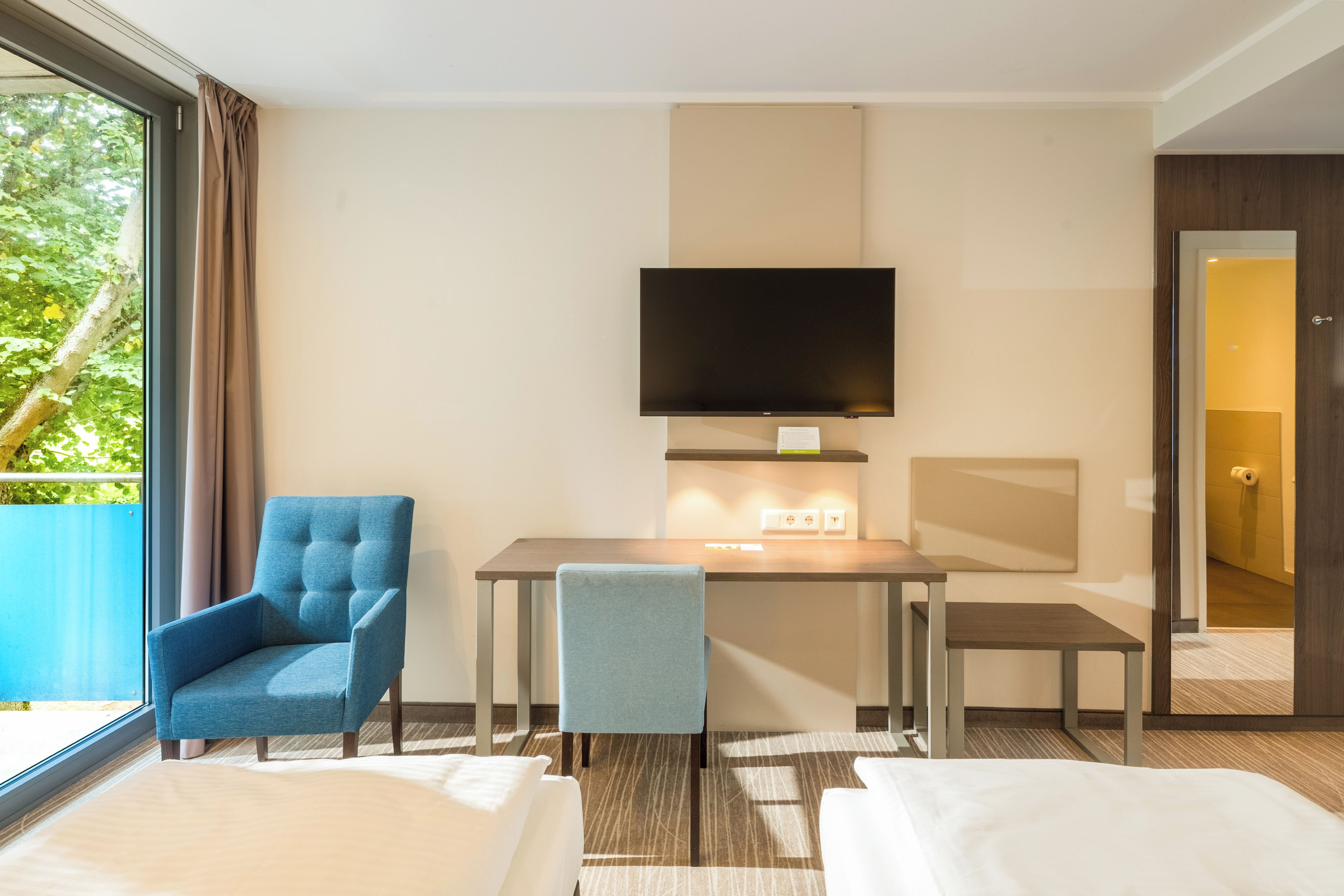 Quarto de hotel com cama, escrivaninha, cadeira, televisão e janela com cortinas.