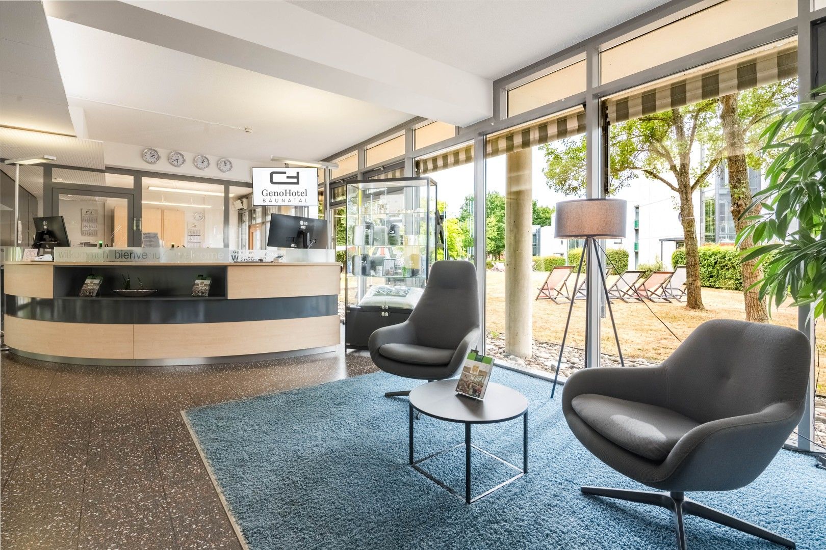 A hotel reception area with a curved desk, two armchairs, a floor lamp, and a view of the outdoor garden.