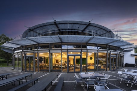 A modern building with a curved glass facade, outdoor seating, and a visible interior with tables and chairs.. GenoHotel Baunatal