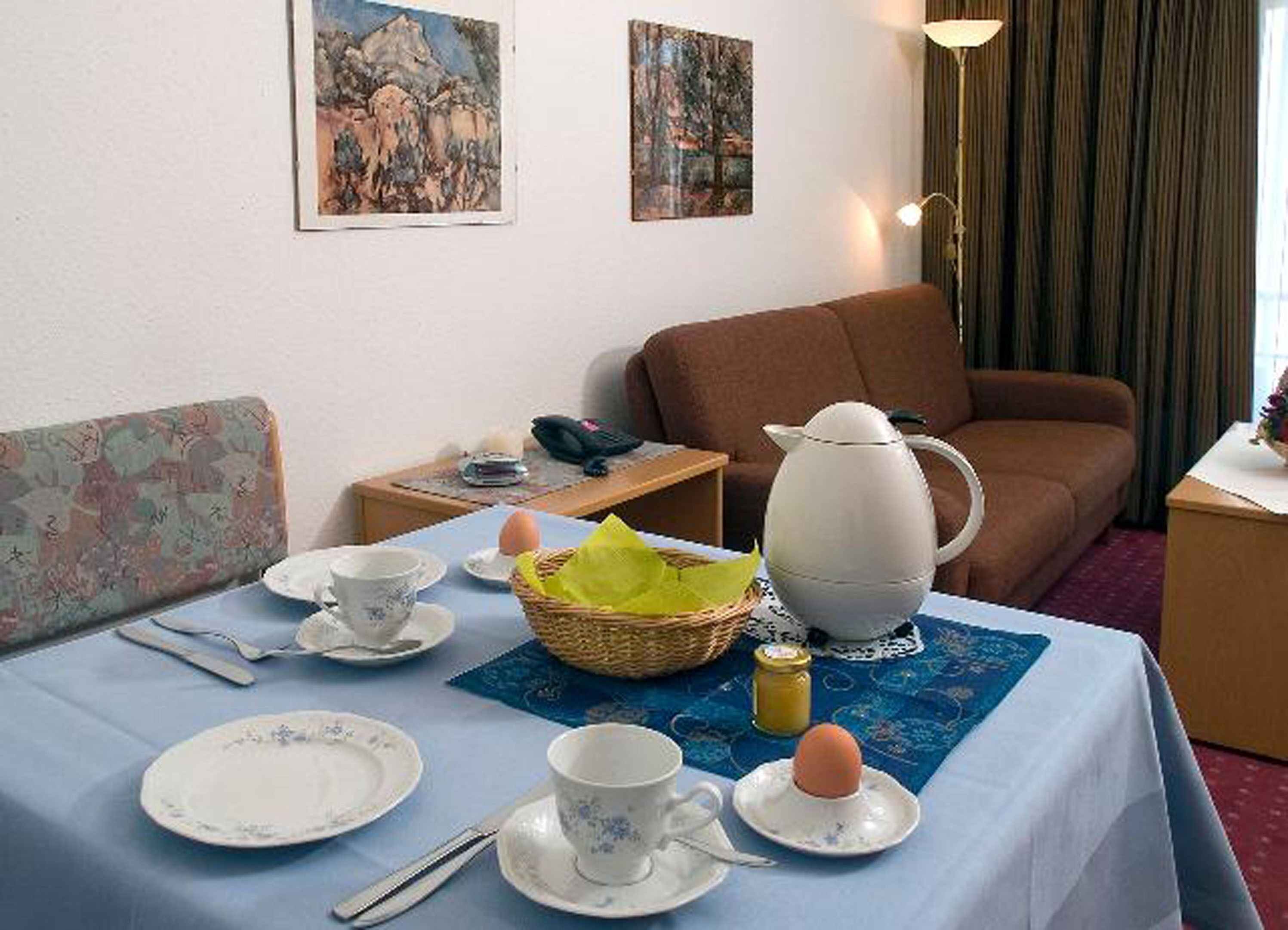 A hotel room with a dining table set for breakfast, a sofa, a lamp, and two paintings on the wall.