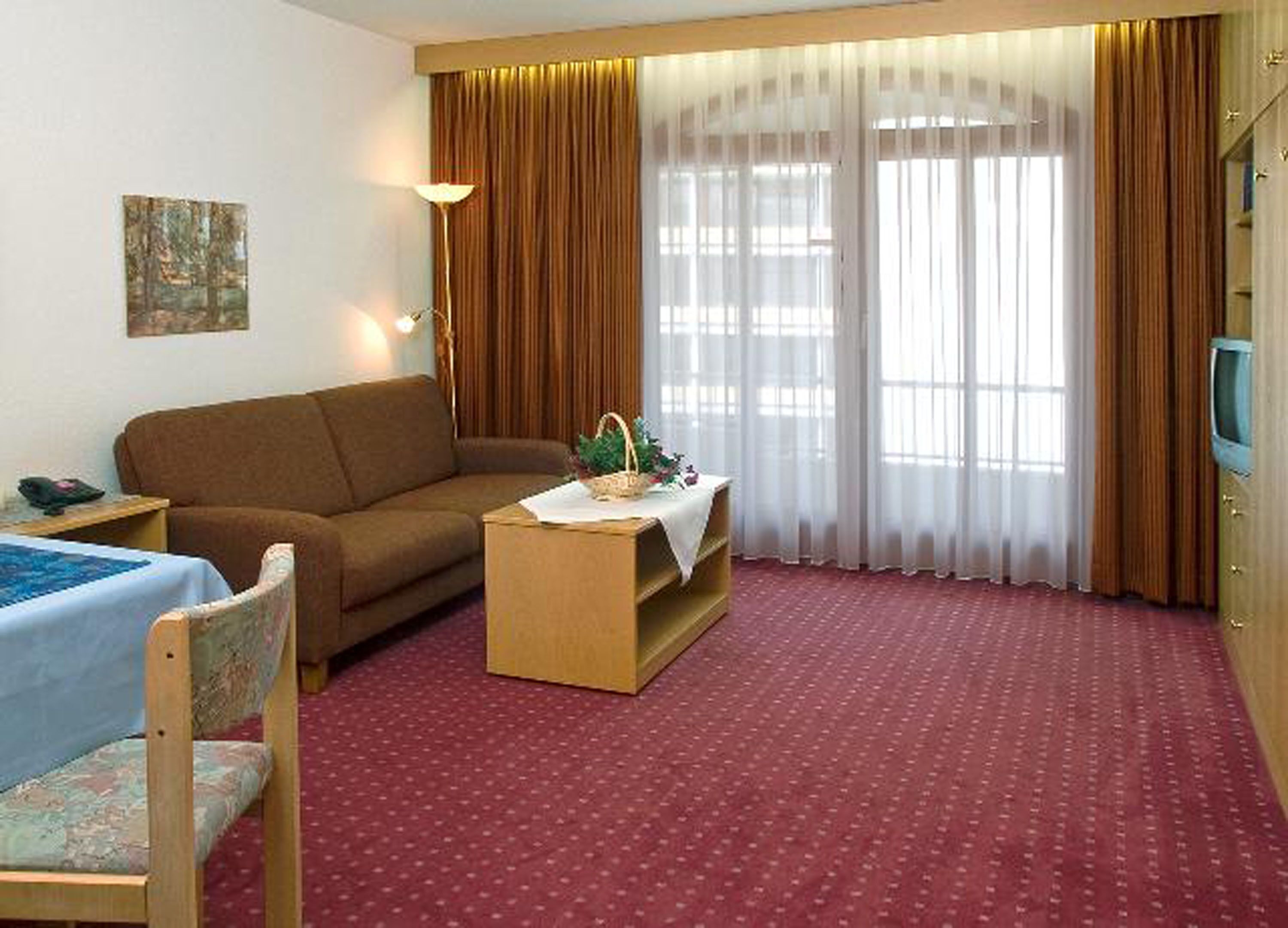 A hotel room with a red carpet, a brown sofa, a wooden coffee table with a plant, a TV on a wooden cabinet, and a painting on the wall.