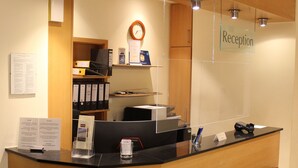 A reception desk with a wooden finish, a clock, and a sign indicating the reception area. - Hotel Bitburger Hof (Bitburg)